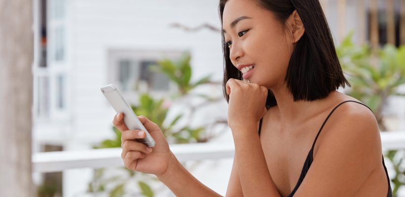 Photo of glad Japanese woman watches funny video on social networks via cell phone, sits in outdoor cafeteria, enjoys chat with boyfriend, texts message, uses free wifi, updates information.