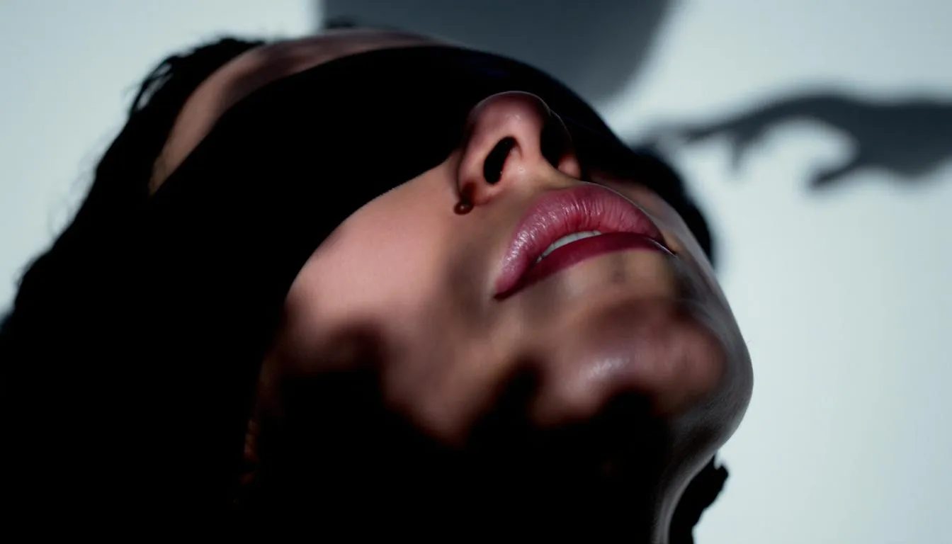 The image depicts a close-up of a person wearing a sleek black blindfold, their head tilted back in anticipation, while a drop of wax hovers above their exposed throat, hinting at the intimate nature of BDSM play. The shadow of their partner looms nearby, emphasizing the power dynamics and sensory exploration involved in this heightened moment of bondage and anticipation.